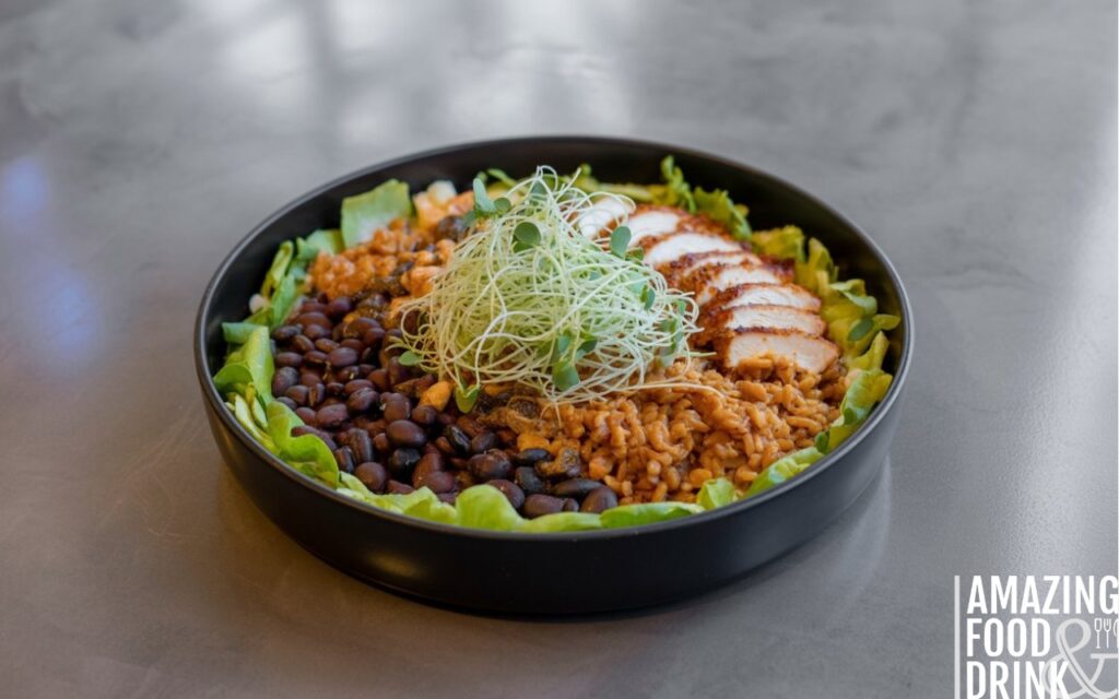 A photograph showcasing an elegant burrito bowl presented with modern plating techniques. The bowl features a vibrant mix of seasoned brown rice, black beans, shredded lettuce, and juicy grilled chicken, all meticulously arranged in a circular pattern within a sleek, matte black bowl. Garnished with delicate microgreens and a sprinkle of toasted pepitas, the arrangement is artfully presented on a polished concrete surface. Soft, diffused lighting highlights the textures and colors of the ingredients, creating a visually appealing and modern culinary composition.