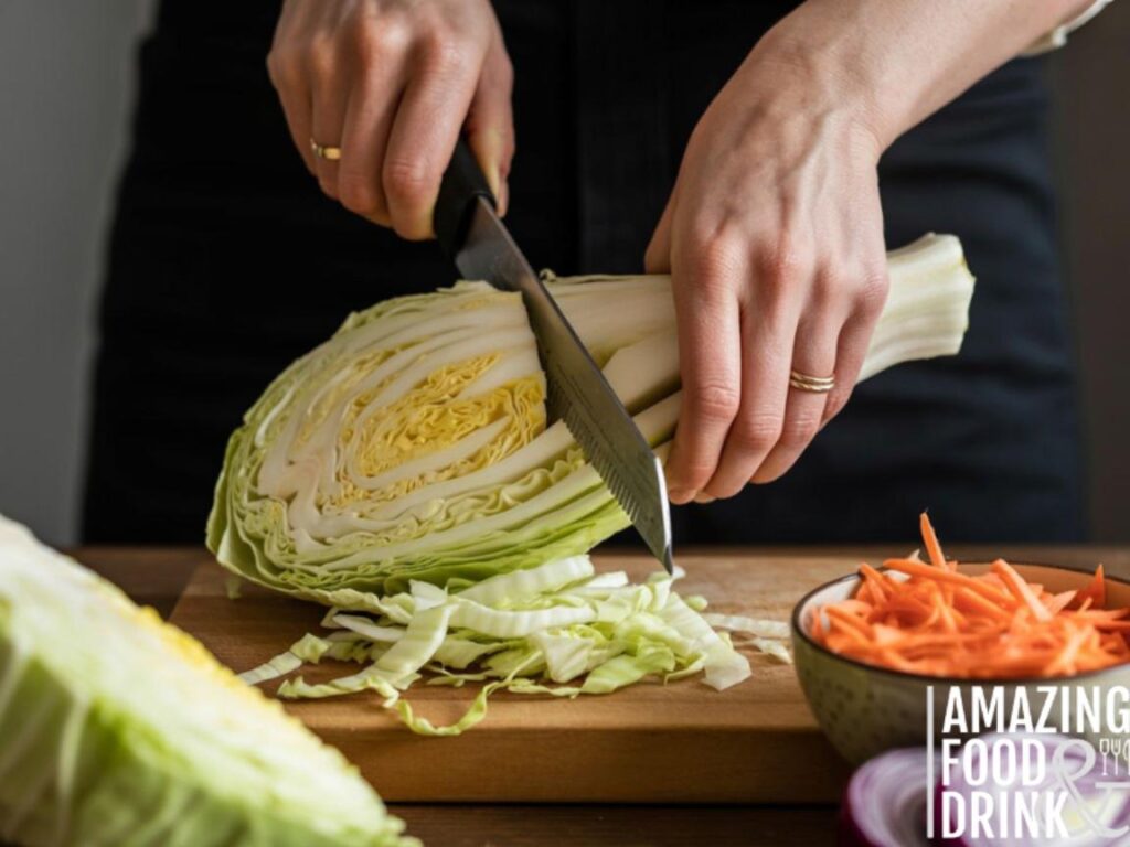 A photograph of hands demonstrating a step-by-step process of shredding cabbage for coleslaw. The hands, adorned with simple gold rings, carefully hold a crisp head of green cabbage, using a mandoline to create thin, even slices that fall onto a wooden cutting board. Beside the cabbage and cutting board sits a small bowl filled with shredded carrots and finely chopped red onion. Soft, natural light illuminates the scene, highlighting the textures of the vegetables and creating a warm, inviting atmosphere.