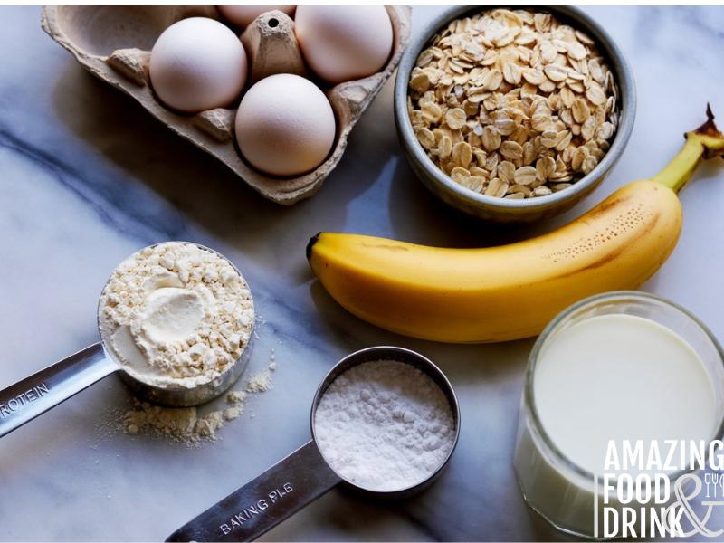 A photograph of a flat lay arrangement showcasing ingredients for protein pancakes on a polished white marble surface. A silver measuring scoop filled with vanilla protein powder sits next to three fresh eggs in a rustic brown carton, alongside a halved ripe banana and a small bowl of rolled oats. A glass of almond milk and a small container of baking powder complete the arrangement, carefully placed to create a balanced and visually appealing composition. Soft, natural light illuminates the scene, highlighting the textures of the ingredients and the cool, smooth surface of the marble.