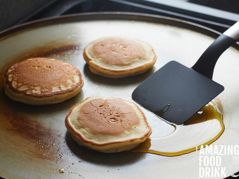 A photograph of protein pancakes cooking on a well-used, silver non-stick griddle. The pancakes are a light golden-brown color, with small bubbles rising to the surface and crispy edges beginning to form. A black silicone spatula rests nearby, poised to flip the pancakes, with a small pool of melted butter shimmering on the griddle. Soft, diffused light illuminates the scene, casting subtle shadows and highlighting the textures of the cooking pancakes.