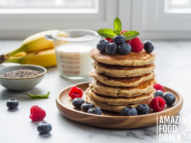 A photograph of a towering stack of protein pancakes artfully arranged on a rustic wooden plate. The pancakes are golden brown with visible texture, and generously garnished with a vibrant assortment of fresh blueberries, raspberries, and a sprig of bright green mint leaves. Surrounding the plate are scattered ingredients: a small bowl of chia seeds, a measuring cup of almond milk, and a few ripe bananas, all bathed in soft, natural window light. A clean white marble countertop forms the background, creating a minimalist and appetizing scene.
