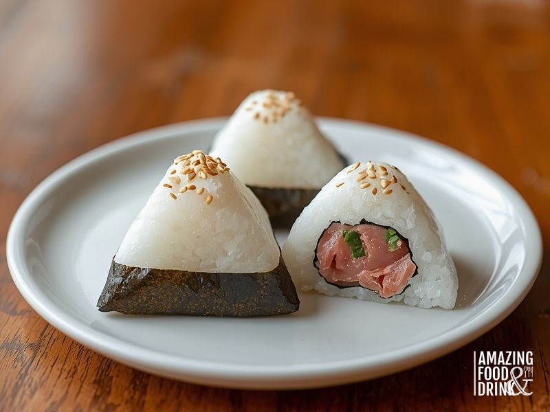 Three onigiri rice balls, prepared with basmati rice, sit on a white plate&mdash;two whole and one cut open to reveal a savory filling of raw fish and green onions for a unique dinner idea.